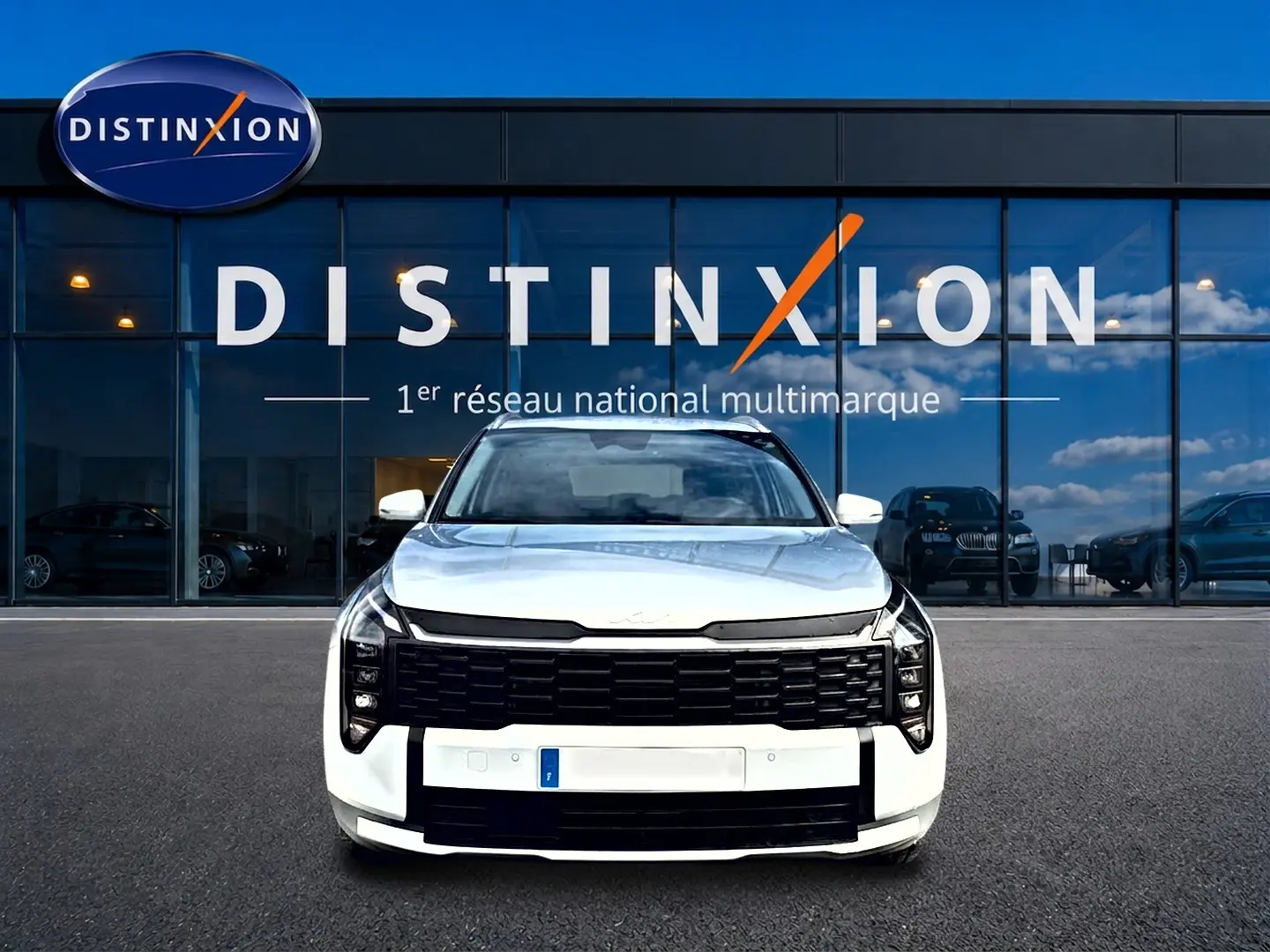 Vue frontale d'un KIA SPORTAGE 1.6 hybride blanc avec calandre noire et phares LED, devant une vitrine Distinxion.