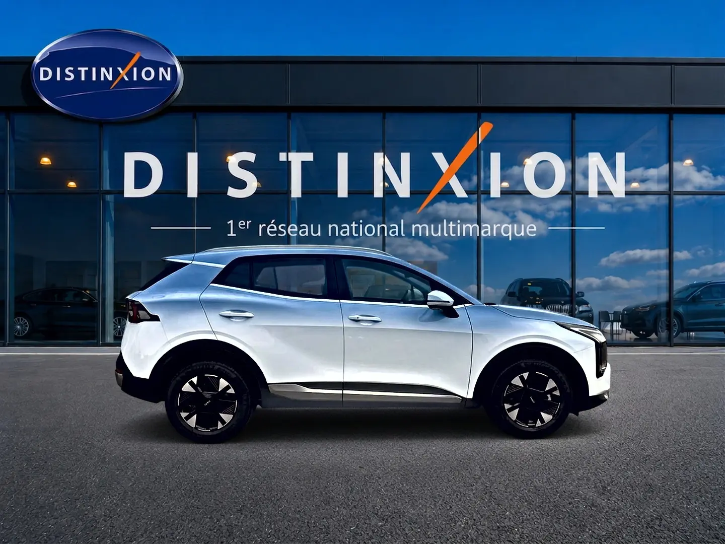 Profil droit d'un KIA SPORTAGE 1.6 hybride 239 blanc, avec jantes alu 17 pouces, devant une vitrine Distinxion.