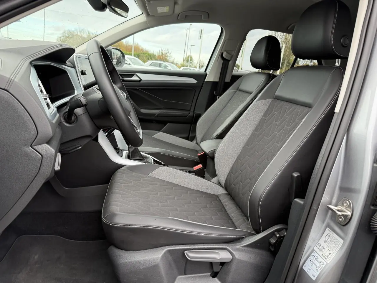 Vue intérieure côté conducteur du Volkswagen T-Roc 2025 argent pyrite, sièges avant en tissu et cuir noir avec surpiqûres blanches.