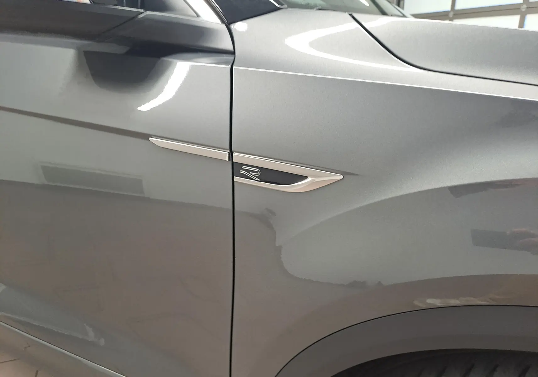 Gros plan sur l'aile avant gauche du Volkswagen T-Roc gris Indium, avec l'emblème R-Line chromé visible.