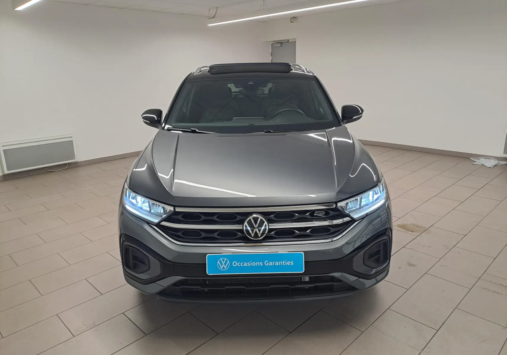 Vue frontale d'un Volkswagen T-Roc 2025 gris Indium avec toit noir et phares LED allumés en intérieur showroom.