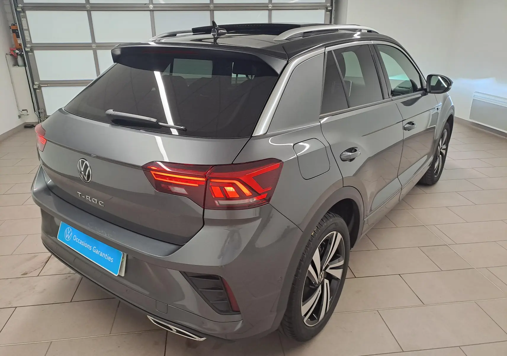 Vue 3/4 arrière droite du Volkswagen T-Roc 2025 gris Indium avec toit noir et feux arrière LED allumés dans un garage.