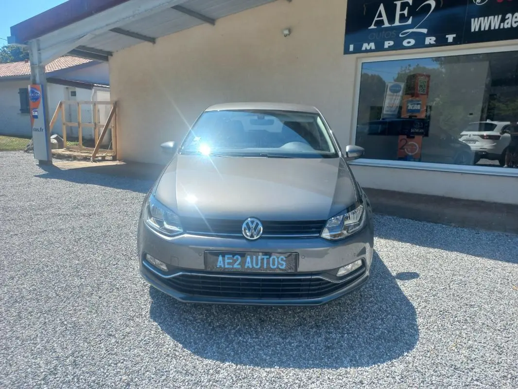 Vue de face d'une Volkswagen Polo gris métallisé 2016, avec plaque AE2 AUTOS, stationnée devant un bâtiment.