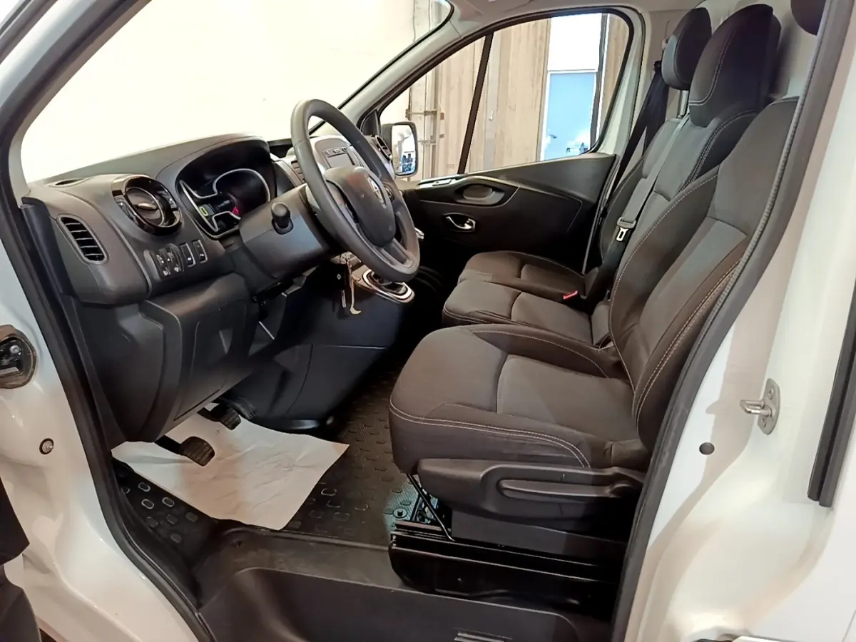 Vue intérieure côté conducteur du Renault Trafic Fourgon blanc, montrant les sièges avant et le tableau de bord noir.