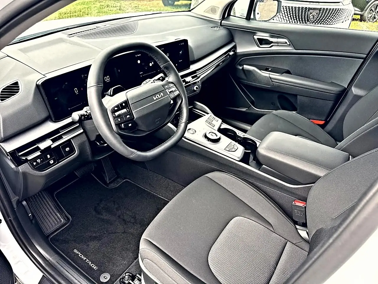 Vue intérieure côté conducteur du Kia Sportage 1.6 hybride blanc, avec volant cuir multifonction et tableau de bord numérique.