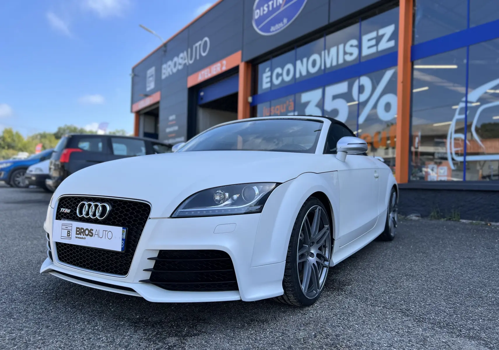 Audi TT RS Roadster blanc vue 3/4 avant avec capote noire, stationné devant un garage Bros Auto.