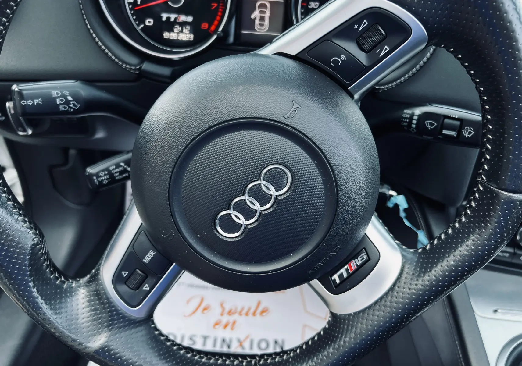 Volant en cuir perforé avec logo Audi au centre, tableau de bord et compte-tours TT RS visibles en arrière-plan.