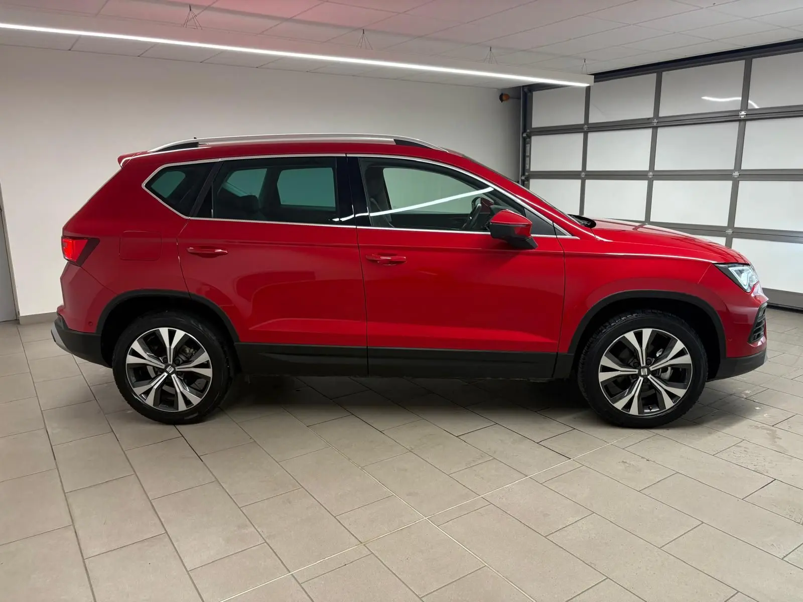 Vue de profil côté gauche d'un SEAT Ateca 2024 Rouge Velvet avec jantes alliage et barres de toit argentées.