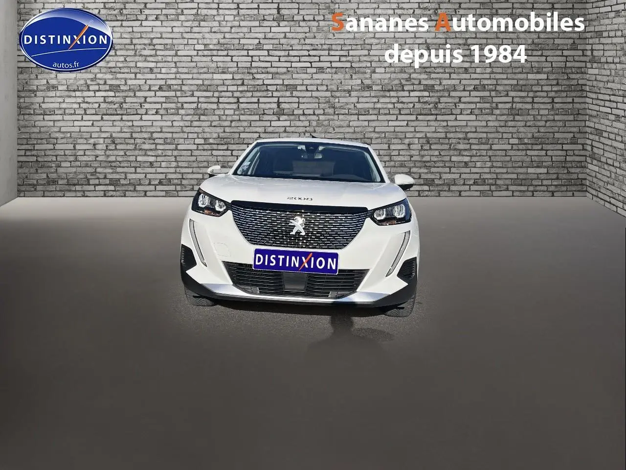 Vue frontale d'un Peugeot 2008 blanc 2020 avec calandre noire et phares LED, sur fond de mur en briques grises.