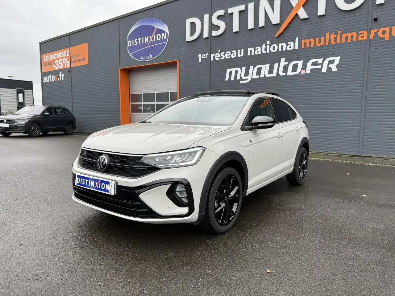 Volkswagen Taigo blanc en 3/4 avant droit avec jantes noires, stationné devant un bâtiment gris et orange.