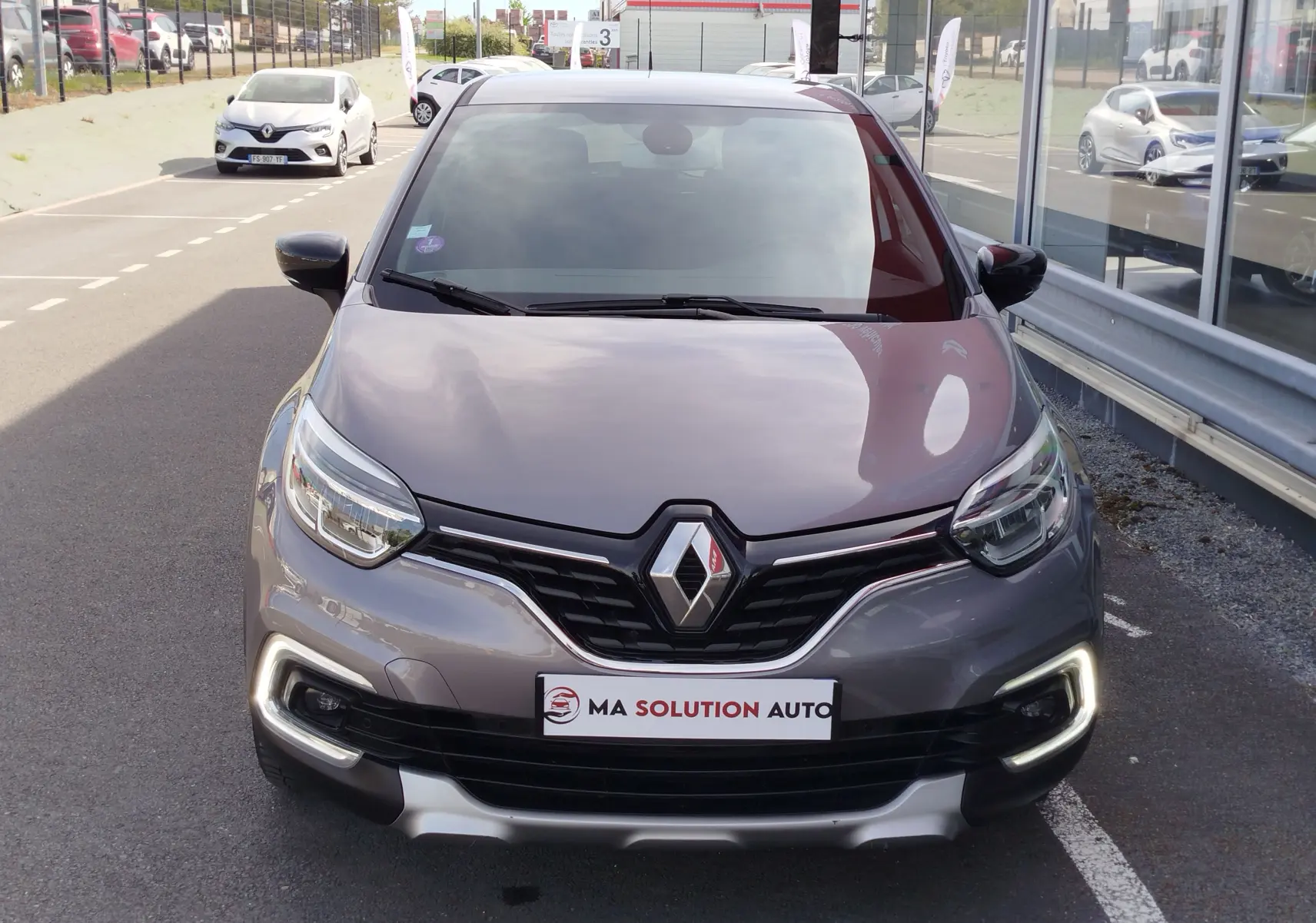 Vue avant d'un Renault Captur gris Cassiopée avec feux diurnes LED allumés, garé devant une vitrine.