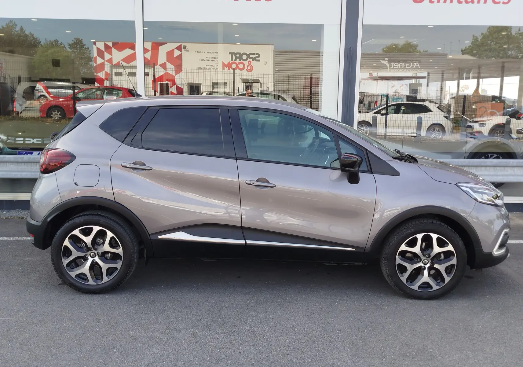 Profil droit d'un Renault Captur gris Cassiopée 2018 avec jantes bi-ton et vitres teintées, stationné devant une vitrine.
