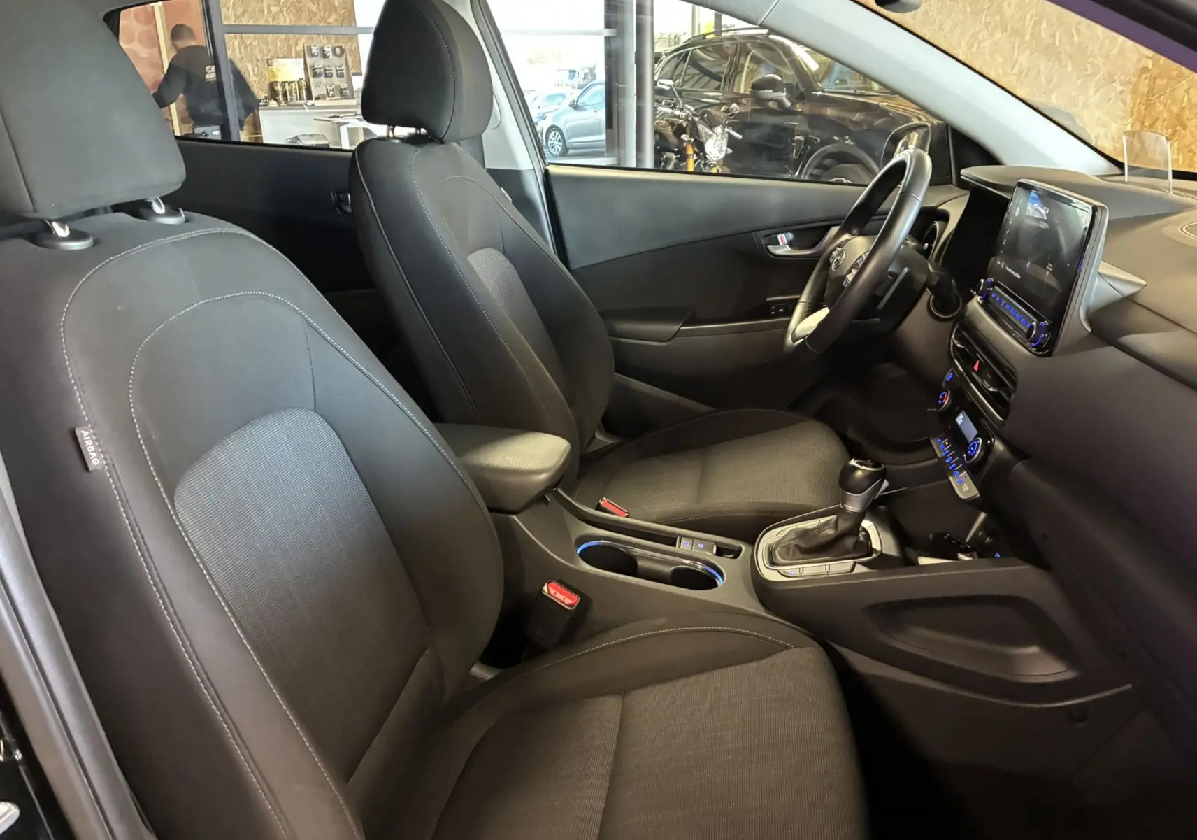 Vue intérieure côté conducteur du Hyundai Kona 2023 noir, sièges tissu noir et tableau de bord avec écran tactile.