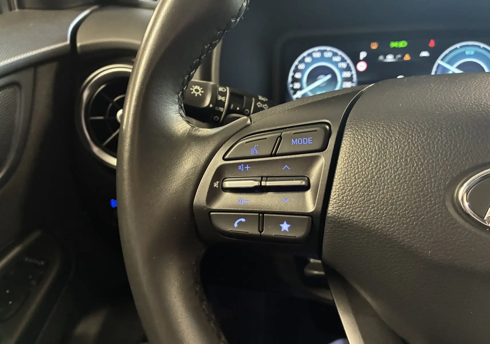 Gros plan sur les commandes gauche du volant noir du Hyundai Kona 2023, avec tableau de bord numérique en arrière-plan.