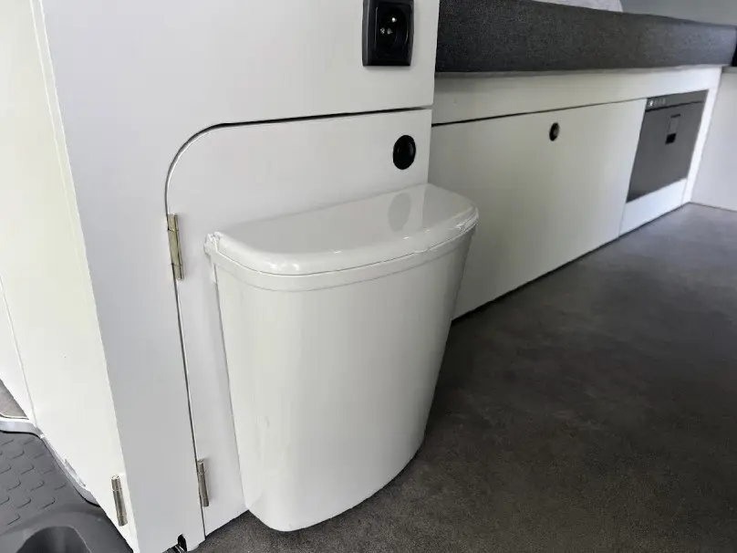 Détail intérieur du fourgon Renault Trafic aménagé, montrant une poubelle blanche intégrée près d'un meuble blanc.