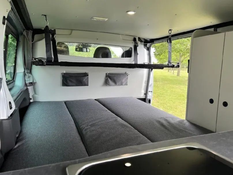 Intérieur du Renault Trafic Fourgon aménagé, vue arrière sur le lit gris et les rangements blancs, avec fenêtre ouverte sur l'extérieur vert.