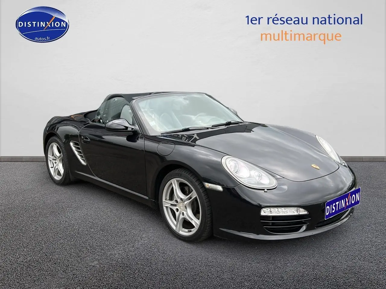 Porsche Boxster 987 noir vue 3/4 avant droit, capote ouverte et jantes alu 17 pouces visibles.