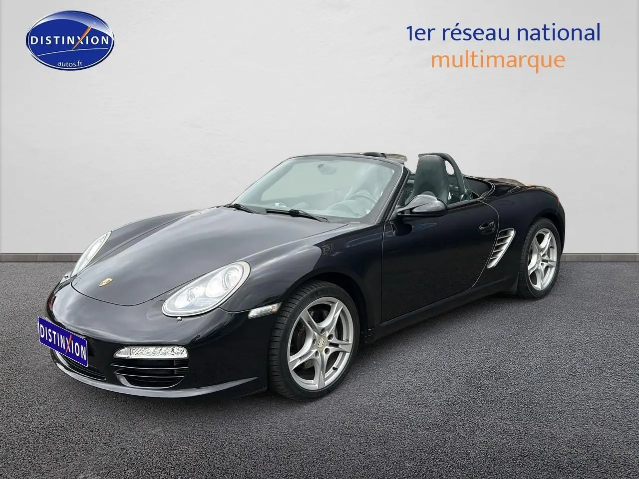 Porsche Boxster (987) noir vue 3/4 avant droit, capote ouverte, jantes alu 17 pouces et phares bi-xénon directionnels.