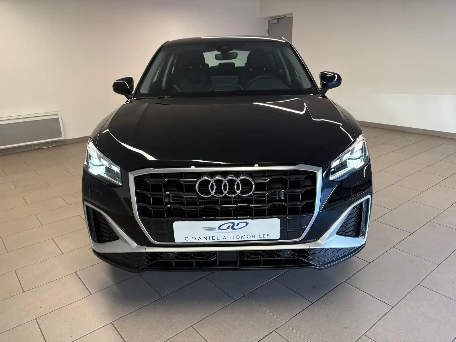 Vue frontale d'un Audi Q2 35 TFSI S Line noir mythic avec calandre hexagonale et phares LED allumés dans un showroom.
