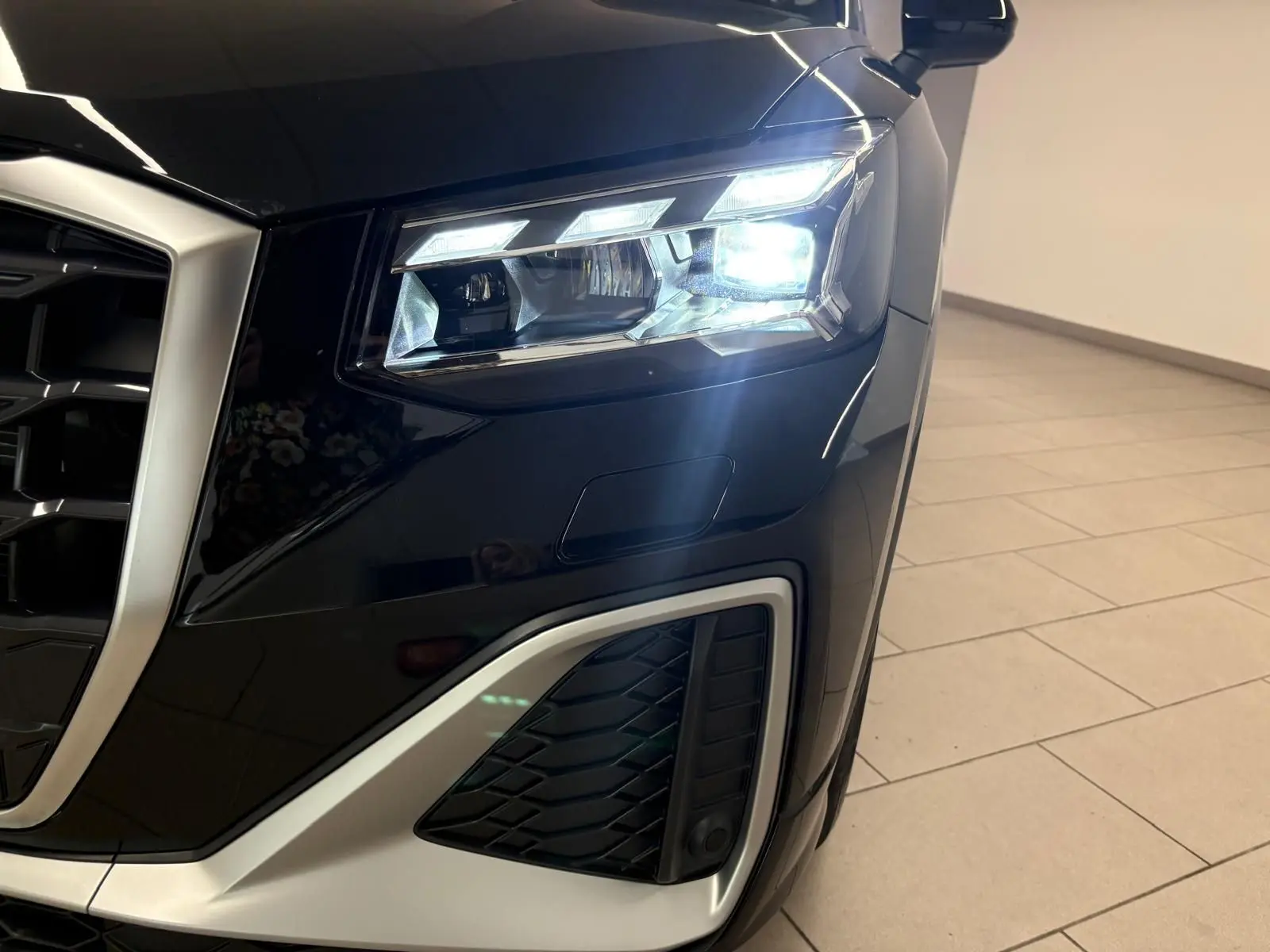 Gros plan sur le phare avant droit allumé de l'Audi Q2 35 TFSI S Line en Noir Mythic, avec calandre et détails argentés visibles.