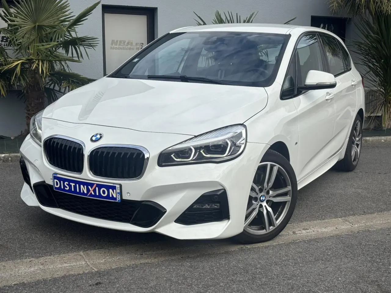 BMW Série 2 Active Tourer blanc vue 3/4 avant droit avec jantes alliage 18 pouces et phares LED allumés.