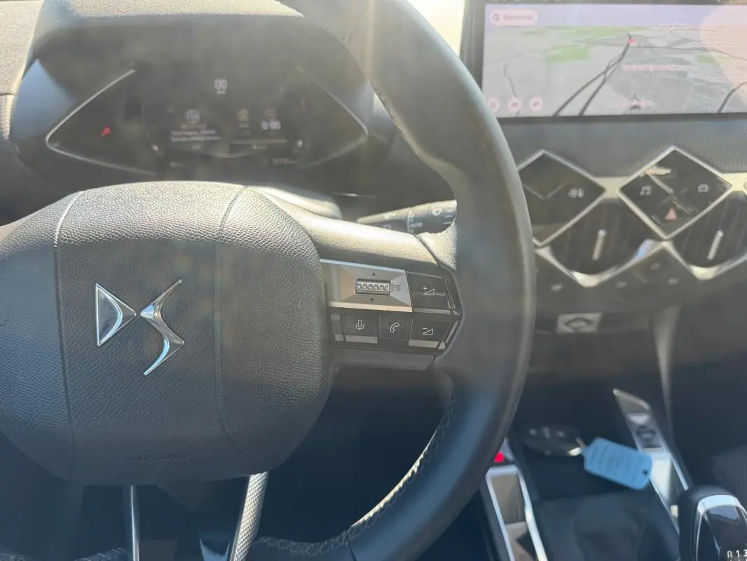 Vue rapprochée du volant noir DS3 2024 avec commandes intégrées et tableau de bord numérique en arrière-plan.