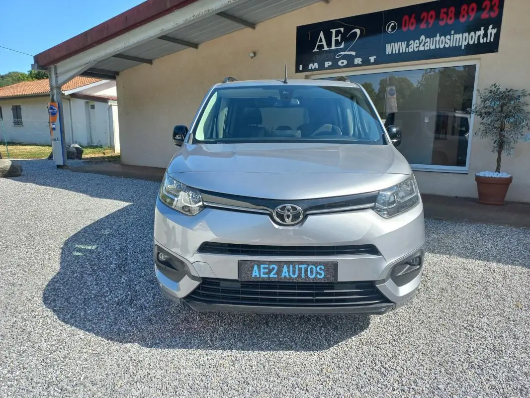 Vue frontale d'un Toyota Proace City Verso gris 2023, avec plaque AE2 AUTOS, stationné devant un bâtiment commercial.