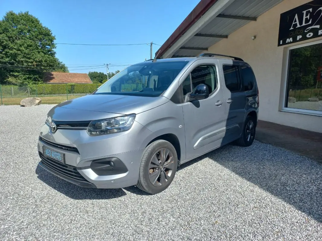 Toyota Proace City Verso gris, vue 3/4 avant droit, avec jantes foncées et toit équipé de barres de toit.