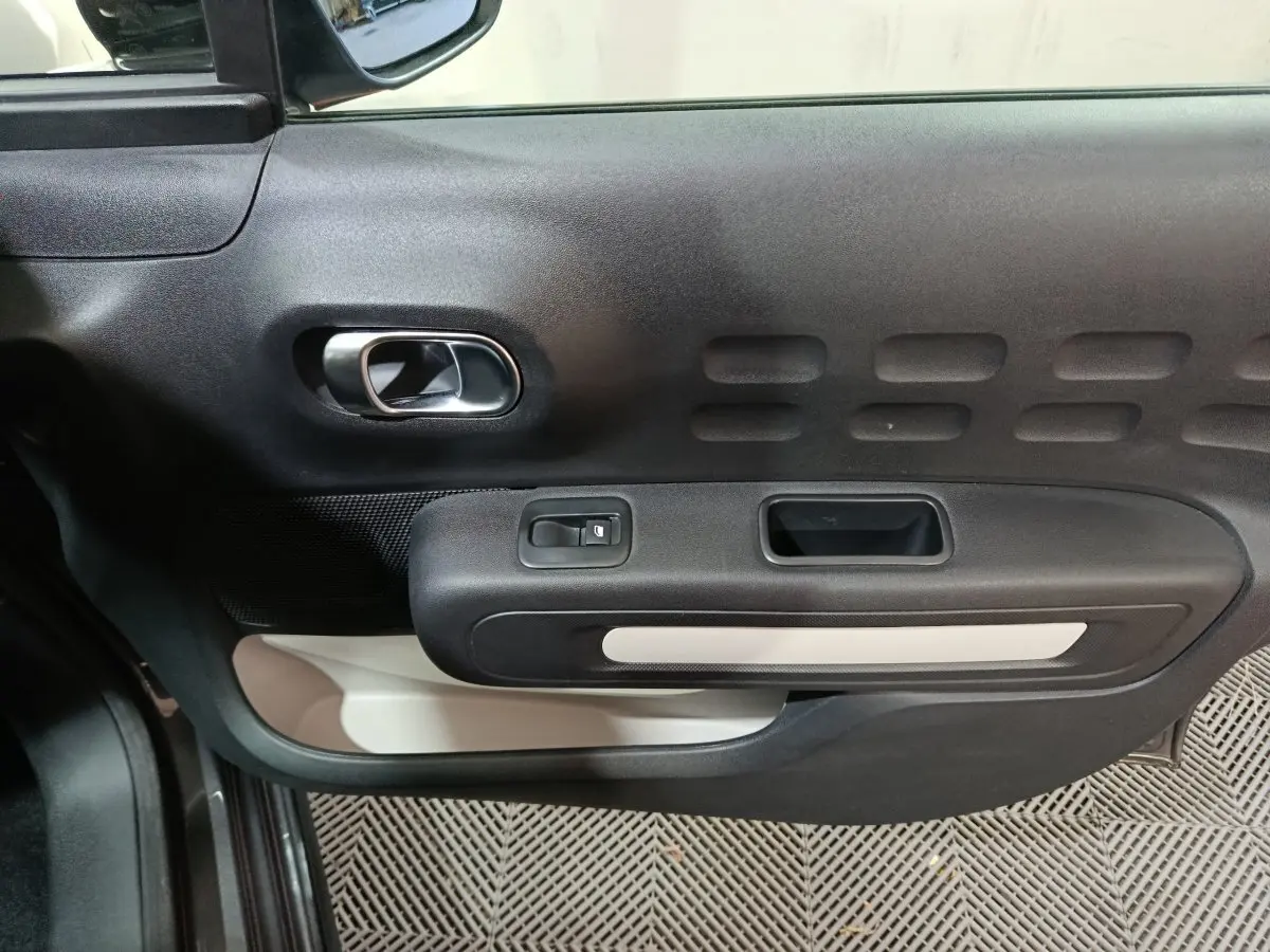 Vue intérieure côté gauche de la porte d’une Citroën C3 2024 gris foncé, avec poignée argentée et commande de vitre.