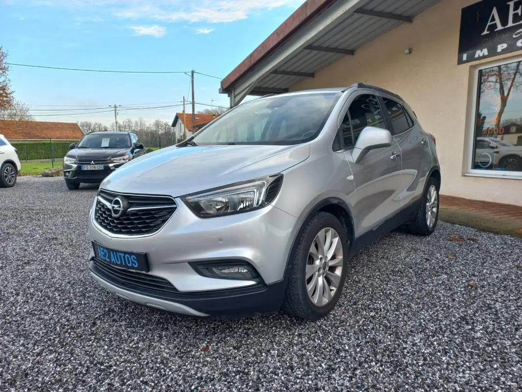 Opel Mokka X gris vue 3/4 avant droit, avec calandre noire et jantes alliage, stationné sur gravier devant un bâtiment.