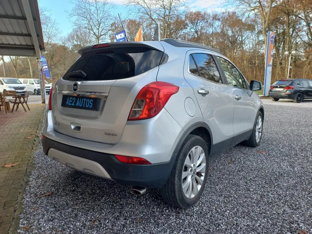 Opel Mokka X gris 2017 vue 3/4 arrière droit, avec feux rouges et jantes alliage visibles, stationné sur gravier.