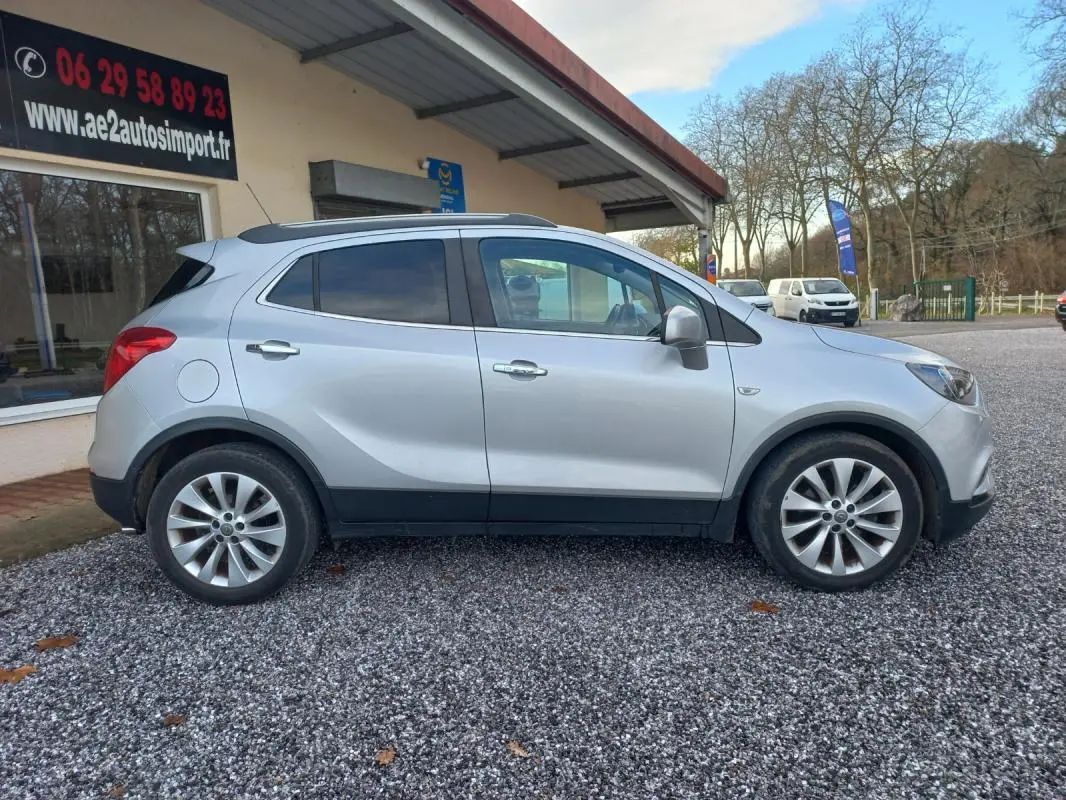 Opel Mokka X gris vue de profil côté gauche, avec jantes alliage et toit ouvrant, stationné devant un bâtiment.