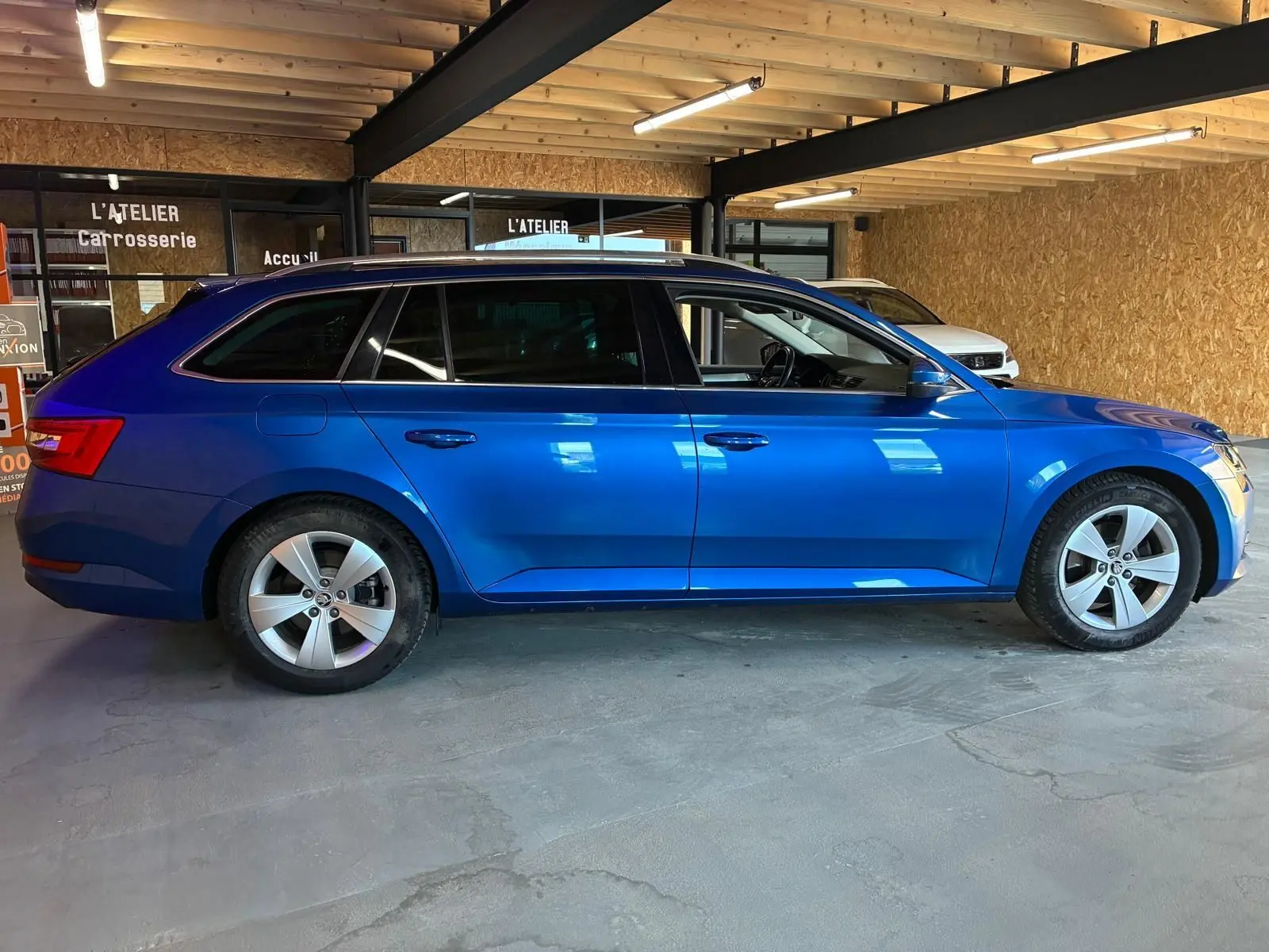 Vue latérale gauche d'une Skoda Superb Combi bleu métallisé 2021 stationnée en intérieur avec jantes alliage 5 branches.