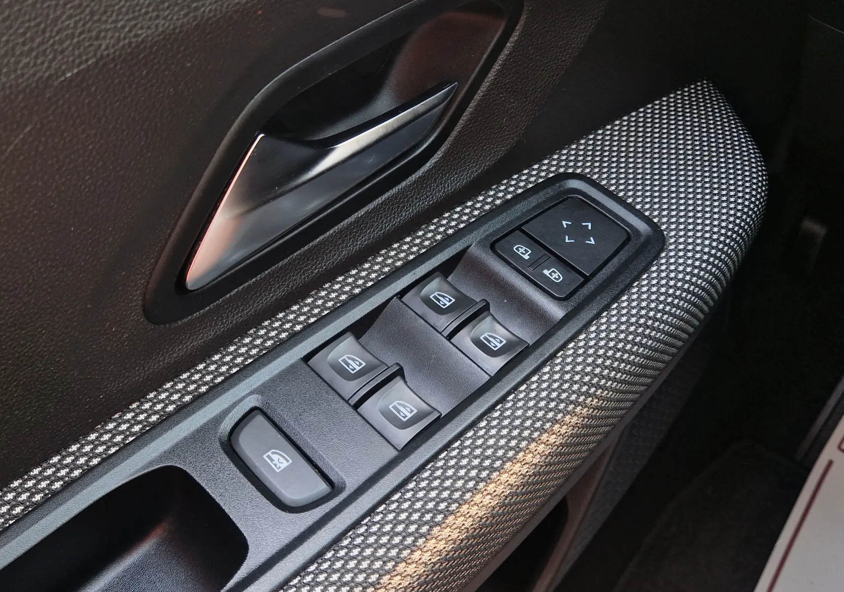 Gros plan sur la commande des vitres électriques côté conducteur d’une Dacia Sandero blanche, tissu gris à motifs.