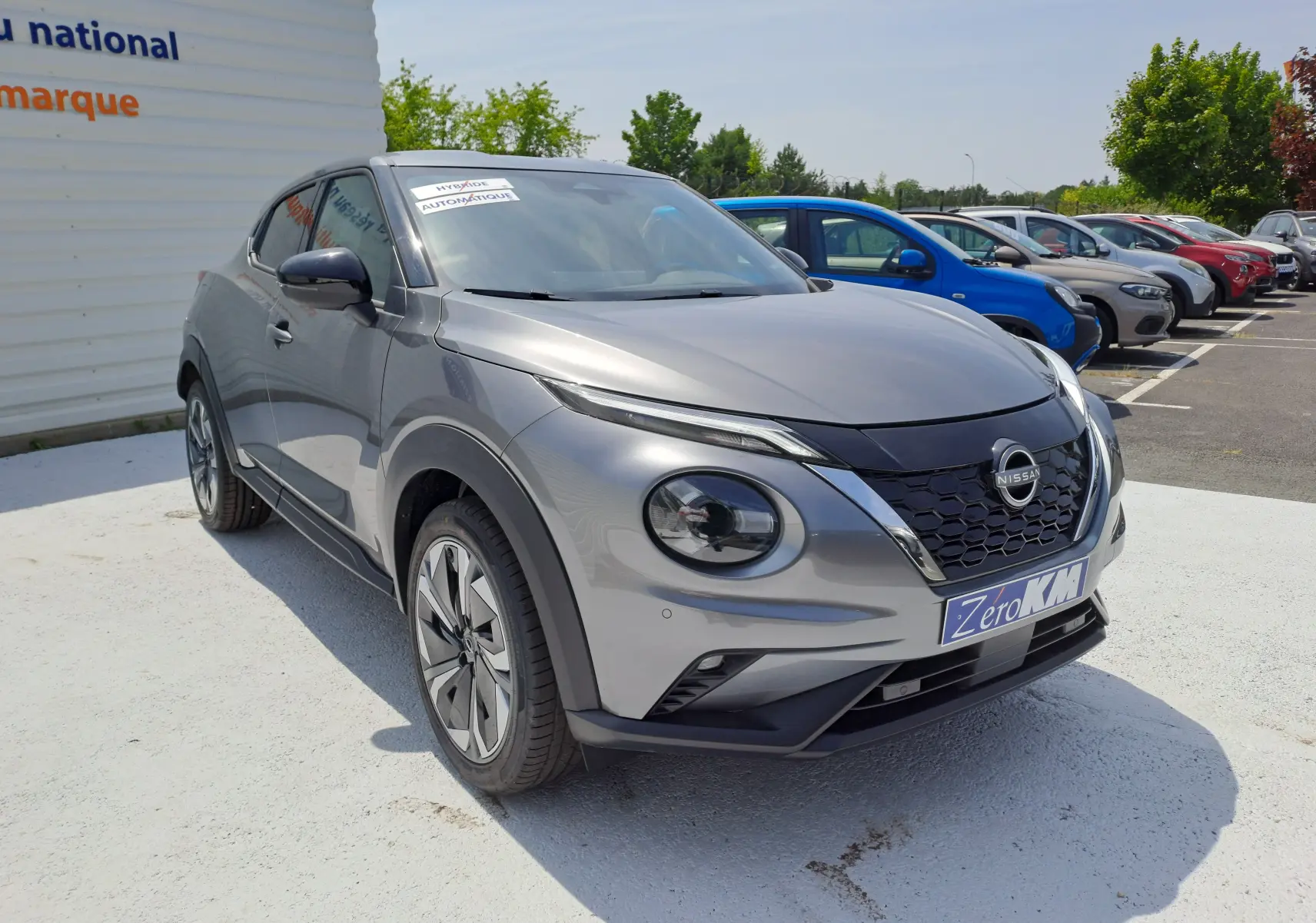 Nissan Juke Hybrid gris squale en 3/4 avant droit, avec calandre noire et jantes modernes visibles.