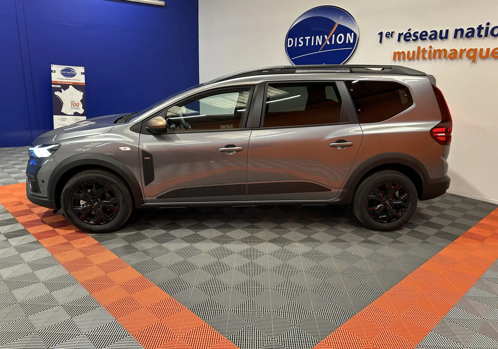 Vue de profil côté gauche d'un Dacia Jogger gris schiste métal avec jantes noires et barres de toit en intérieur showroom.