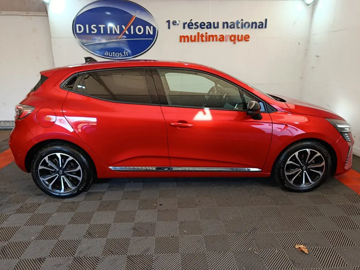 Profil droit d'une Renault Clio Techno TCe 90 rouge 2024 avec jantes alliage et vitres teintées en intérieur showroom.