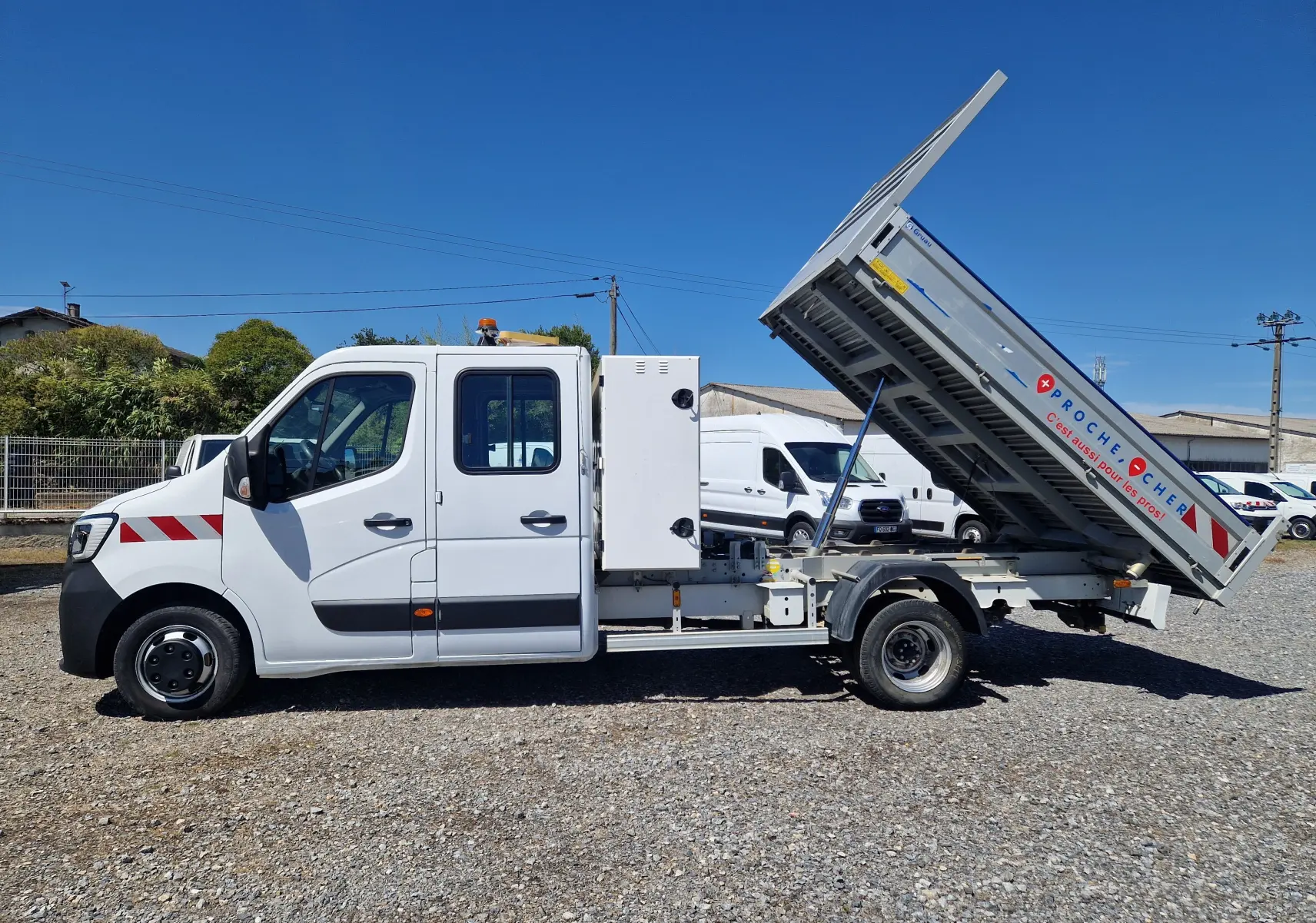 Profil côté gauche d’un Renault Master blanc avec benne relevée et coffre latéral visible sur un sol gravillonné.