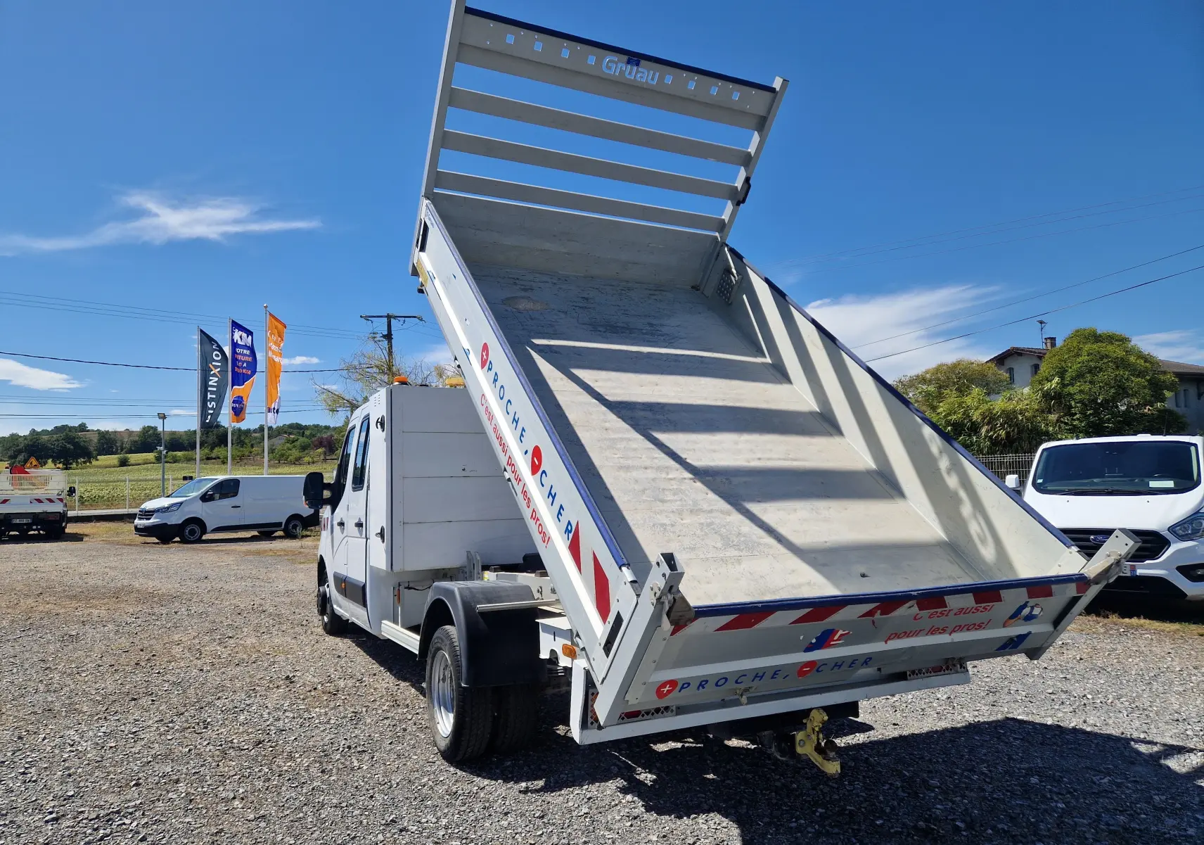 Vue 3/4 arrière droite du Renault Master blanc avec benne relevée et coffre latéral visible sur un terrain ensoleillé.