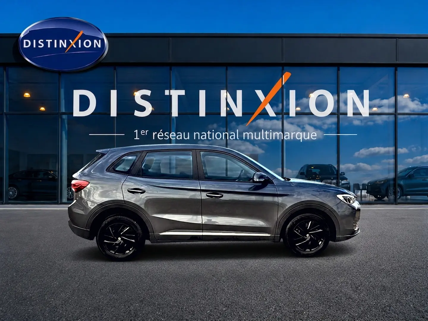 Profil droit du MG ZS Camden grey hybride 2026, avec jantes noires et lignes épurées devant un showroom Distinxion.
