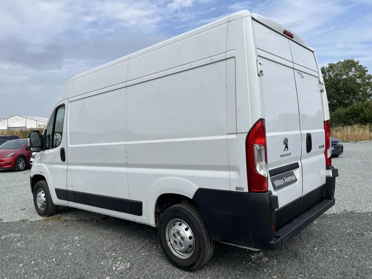 Vue 3/4 arrière droite d'un Peugeot Boxer fourgon blanc avec portes arrière battantes et protections noires sur un sol gravillonné.
