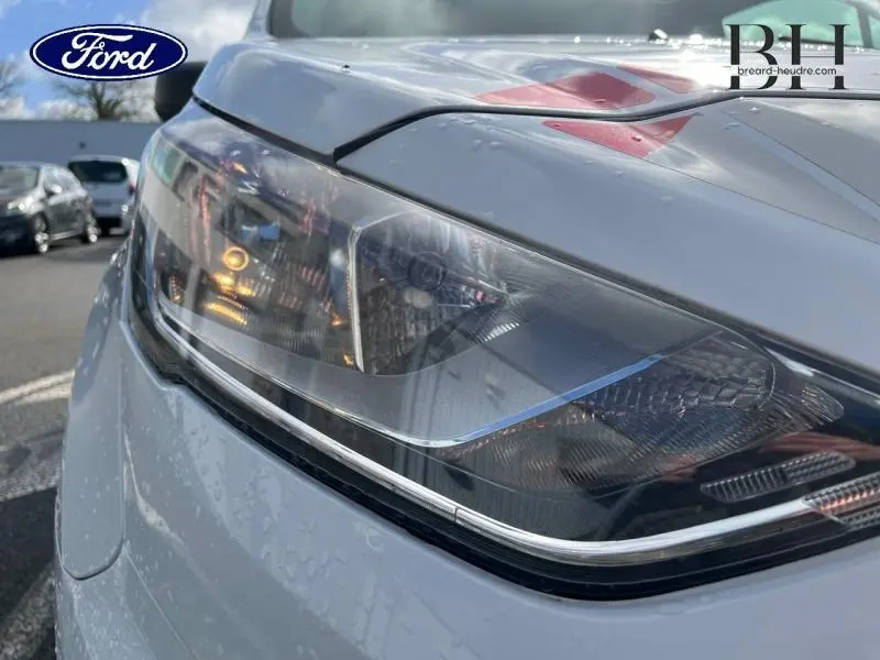 Gros plan sur le phare avant droit du Ford Transit Connect blanc glacier, avec reflets et gouttes d'eau visibles.