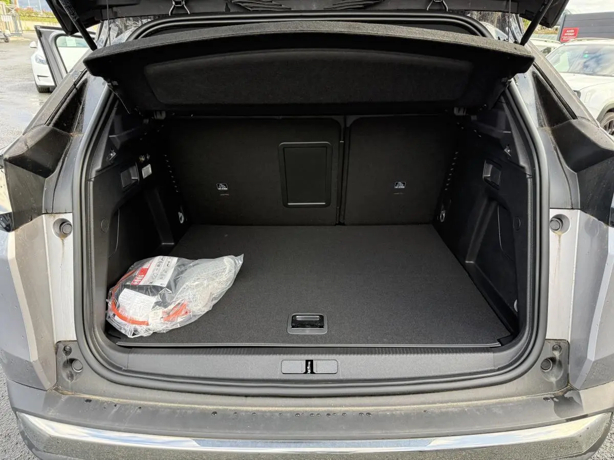 Coffre ouvert vu de l'arrière du Peugeot 3008 Hybrid gris, avec tapis noir et kit de dépannage visible à gauche.