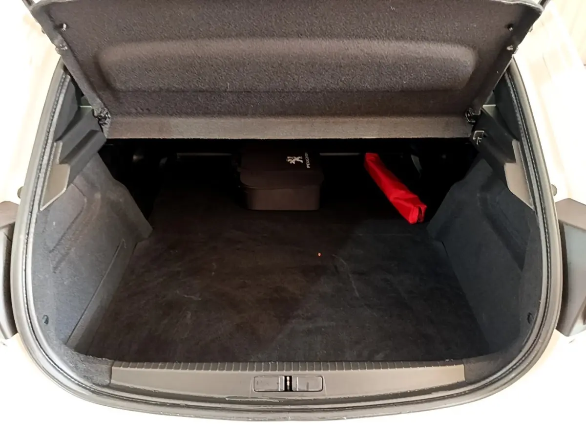 Vue intérieure du coffre ouvert d'une Peugeot 208 électrique blanche, avec tapis noir et accessoires rangés à l'arrière.