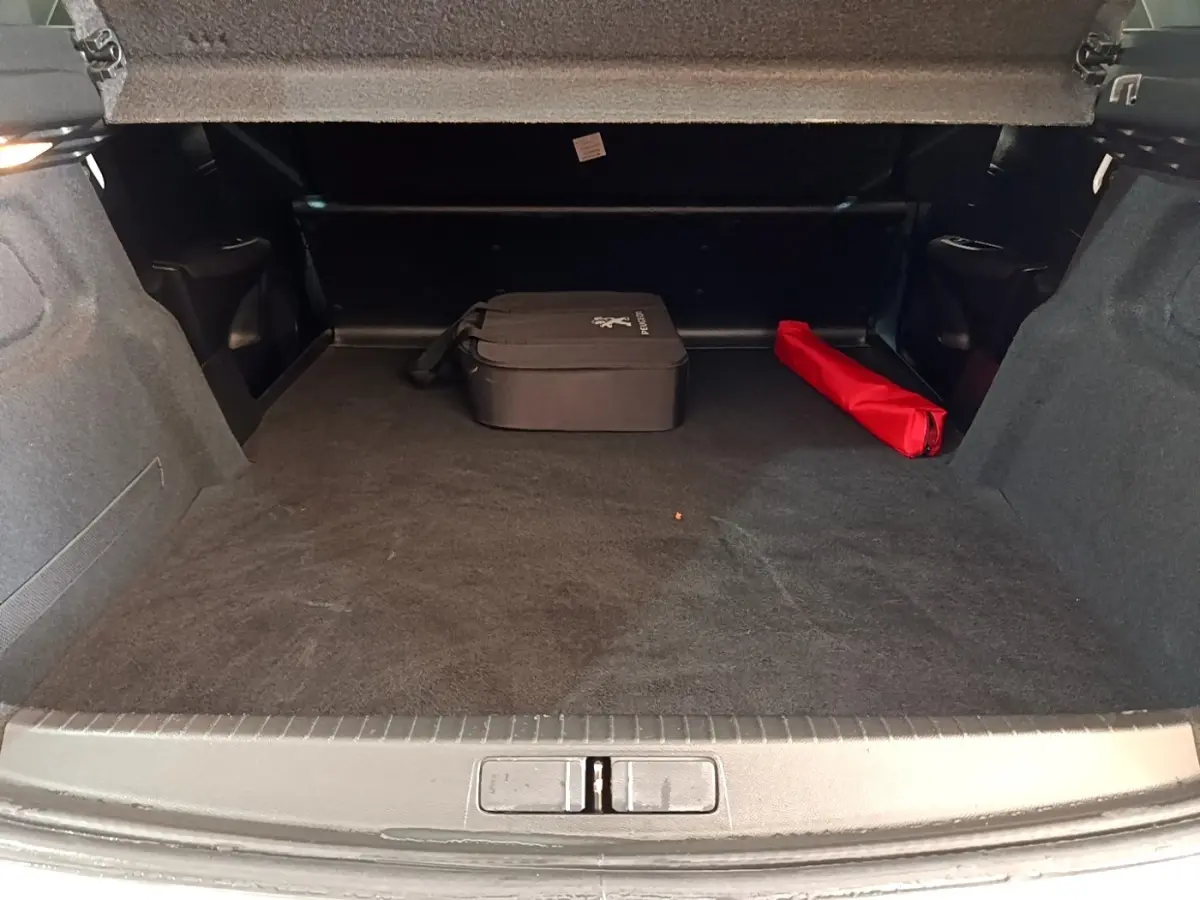 Coffre ouvert d’une Peugeot 208 électrique blanche, montrant un espace de chargement avec une trousse noire et un objet rouge.
