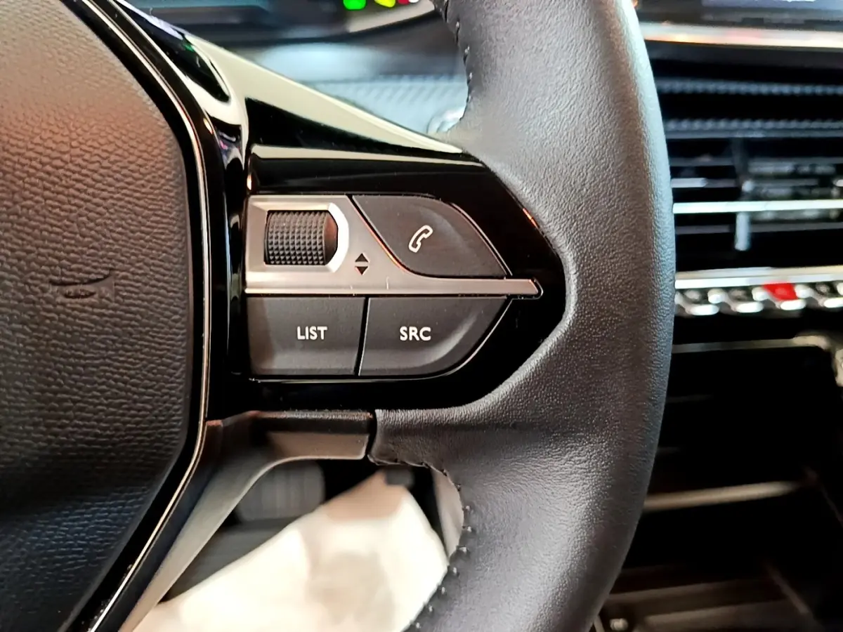 Gros plan sur les commandes gauche du volant cuir noir du Peugeot 208 électrique blanc, avec boutons de gestion audio et appel.