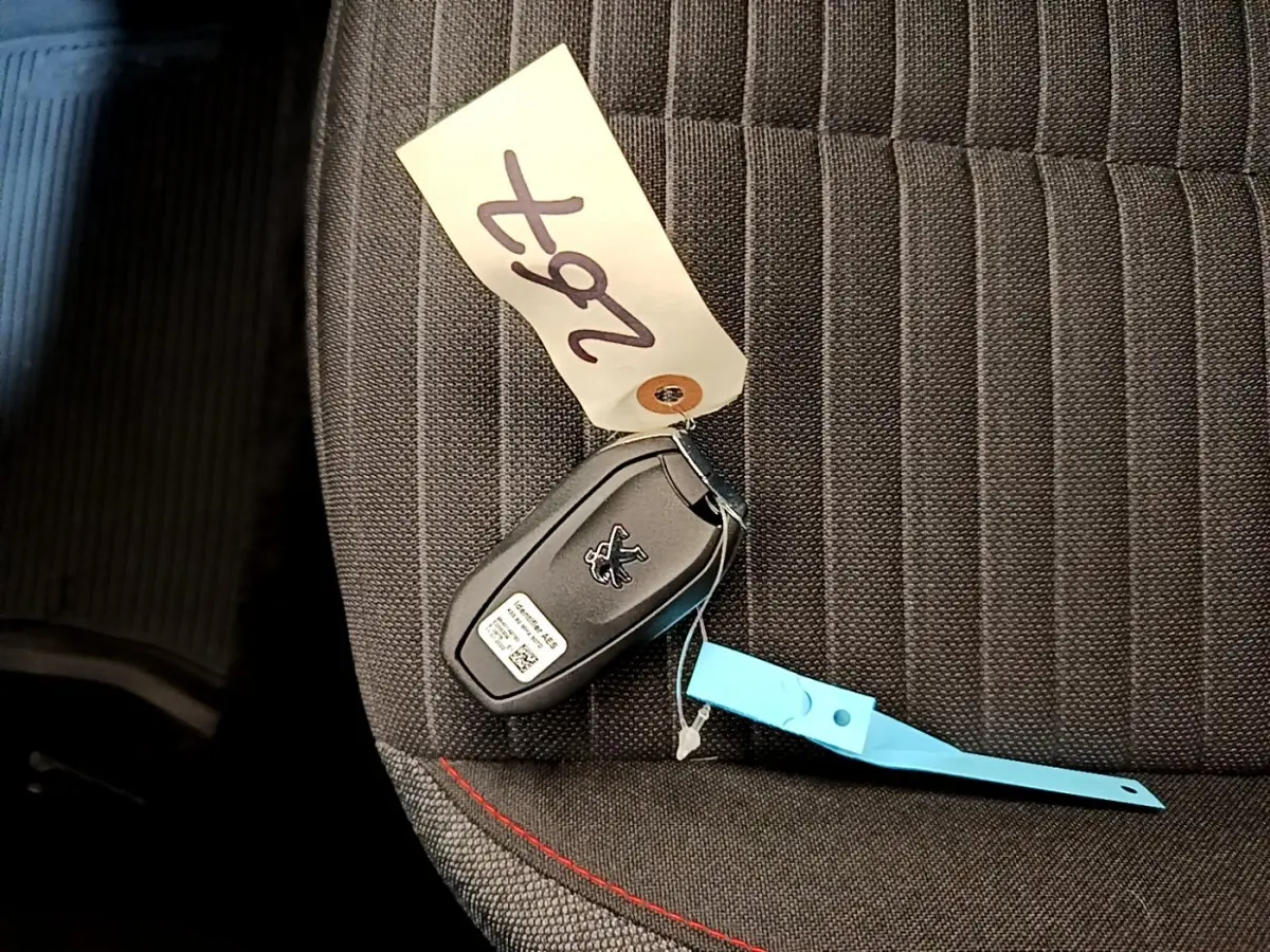 Clé de voiture Peugeot posée sur un siège tissu gris foncé avec surpiqûres rouges, intérieur visible partiellement.
