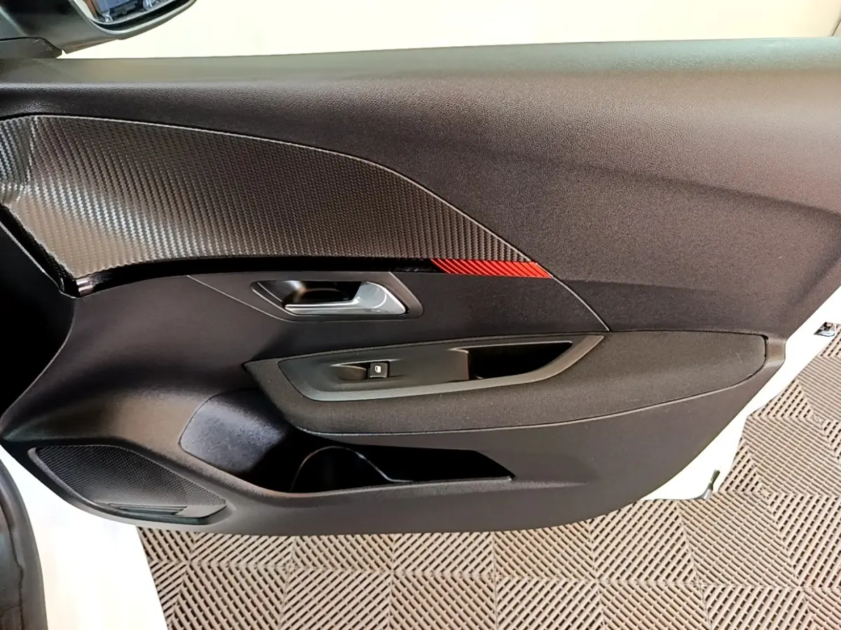 Vue intérieure du panneau de porte côté conducteur de la Peugeot 208 électrique blanche, avec insert carbone et détail rouge.