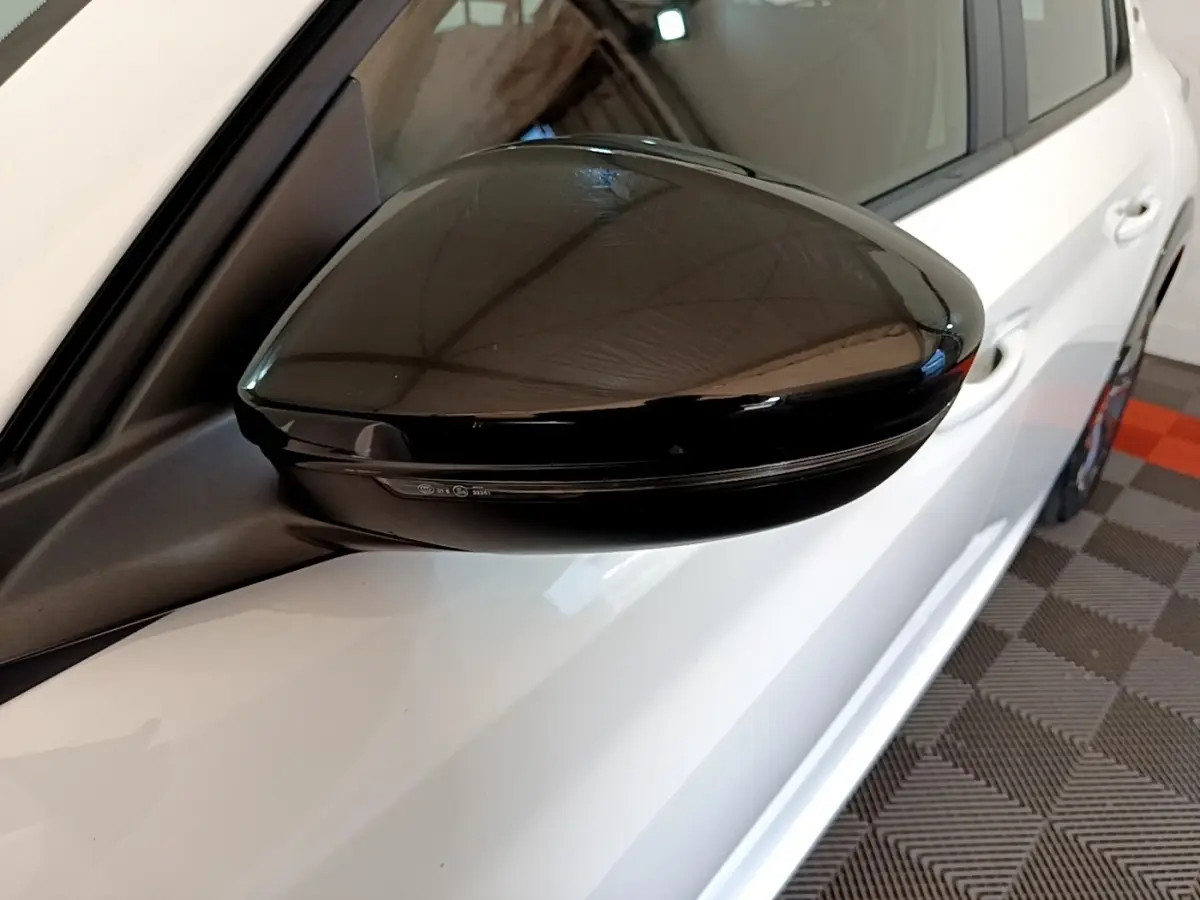 Rétroviseur noir côté gauche en gros plan sur Peugeot 208 électrique blanche vue partielle côté conducteur.