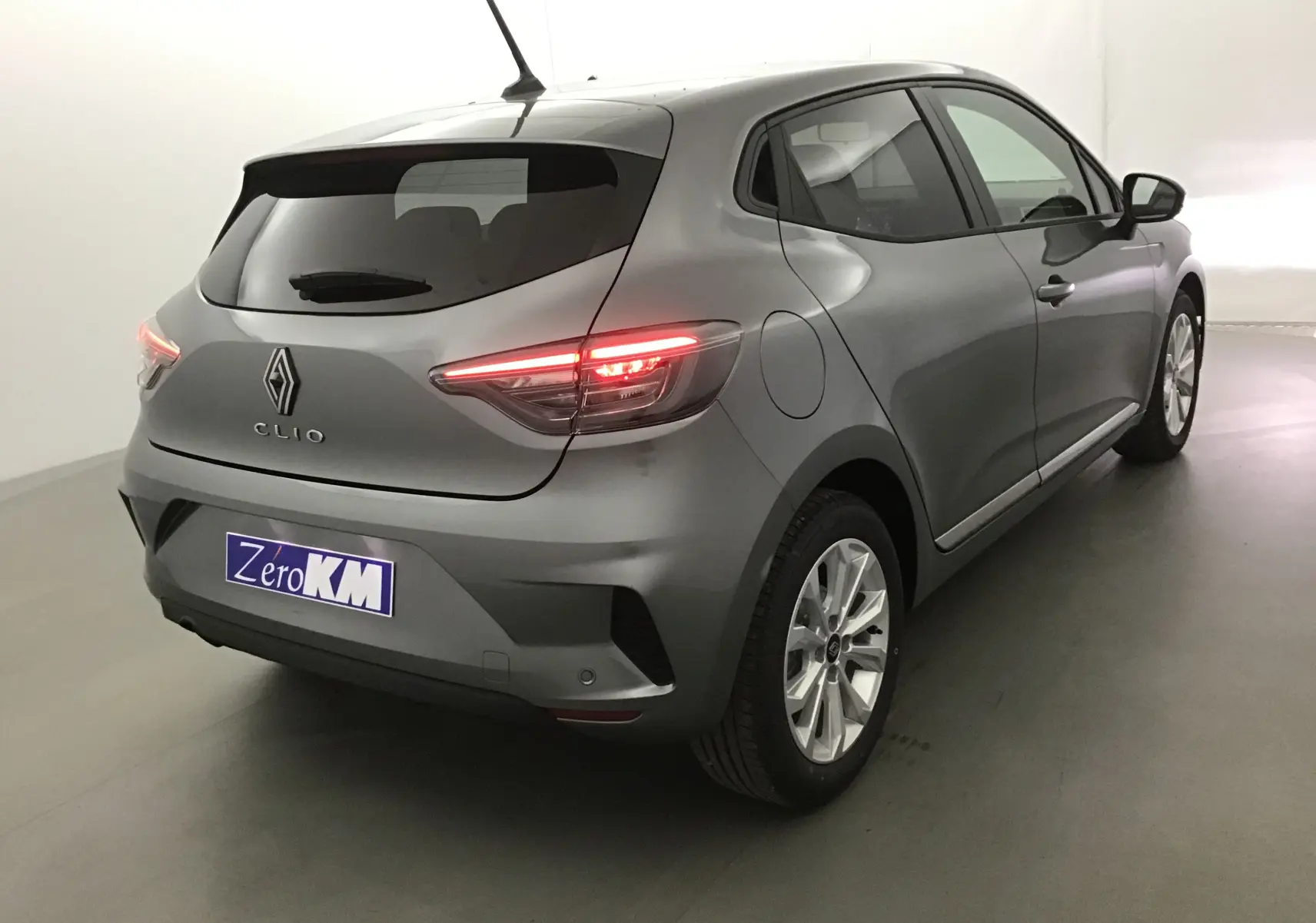 Vue 3/4 arrière droite d'une Renault Clio V gris schiste 2025 avec feux allumés et protections latérales chromées visibles.